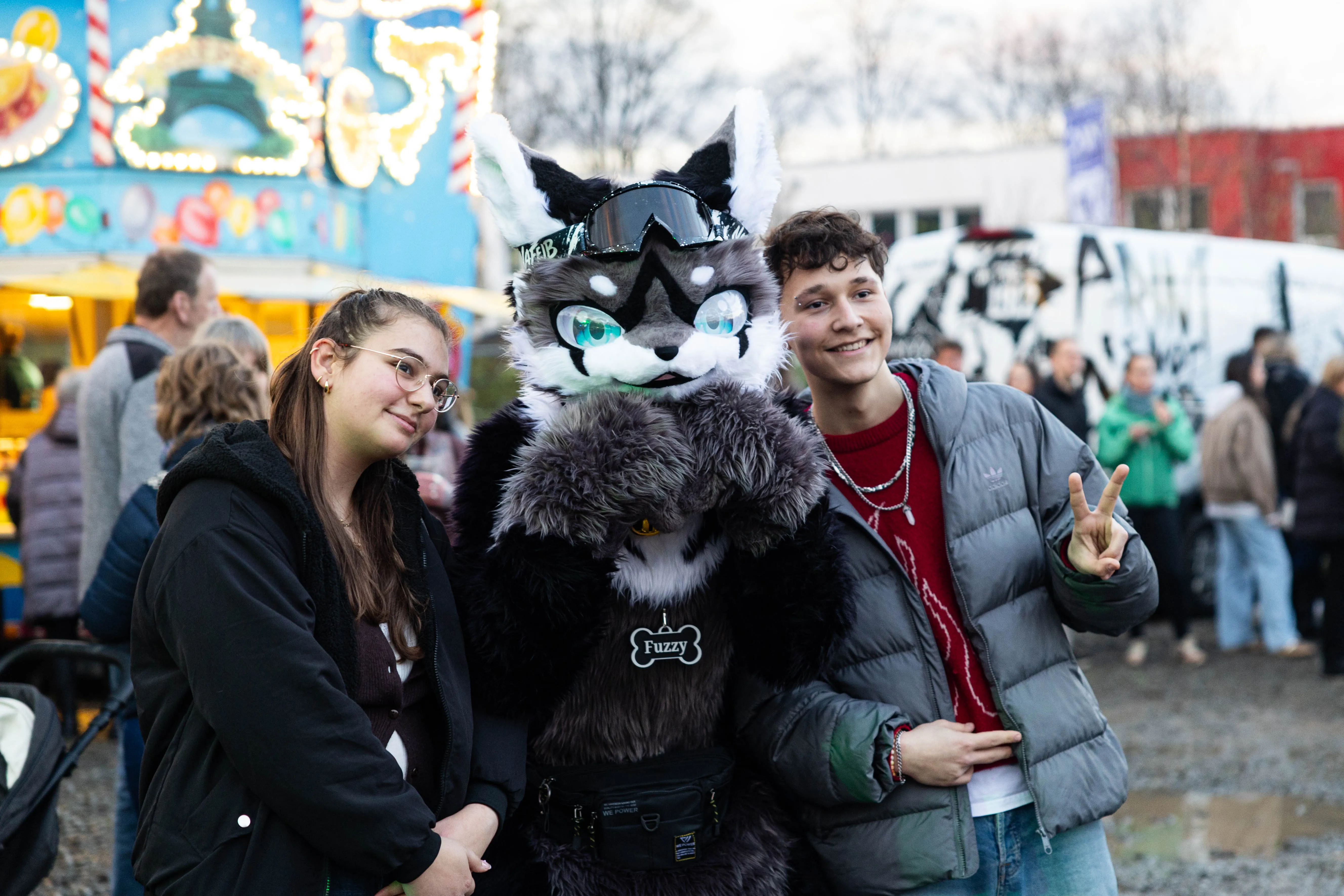 Zwei junge Erwachsene stehen neben einer Person in einem aufwendigen Fuchs-Kostüm auf einem Volksfest. Im Hintergrund sind Fahrgeschäfte und Menschen zu sehen.