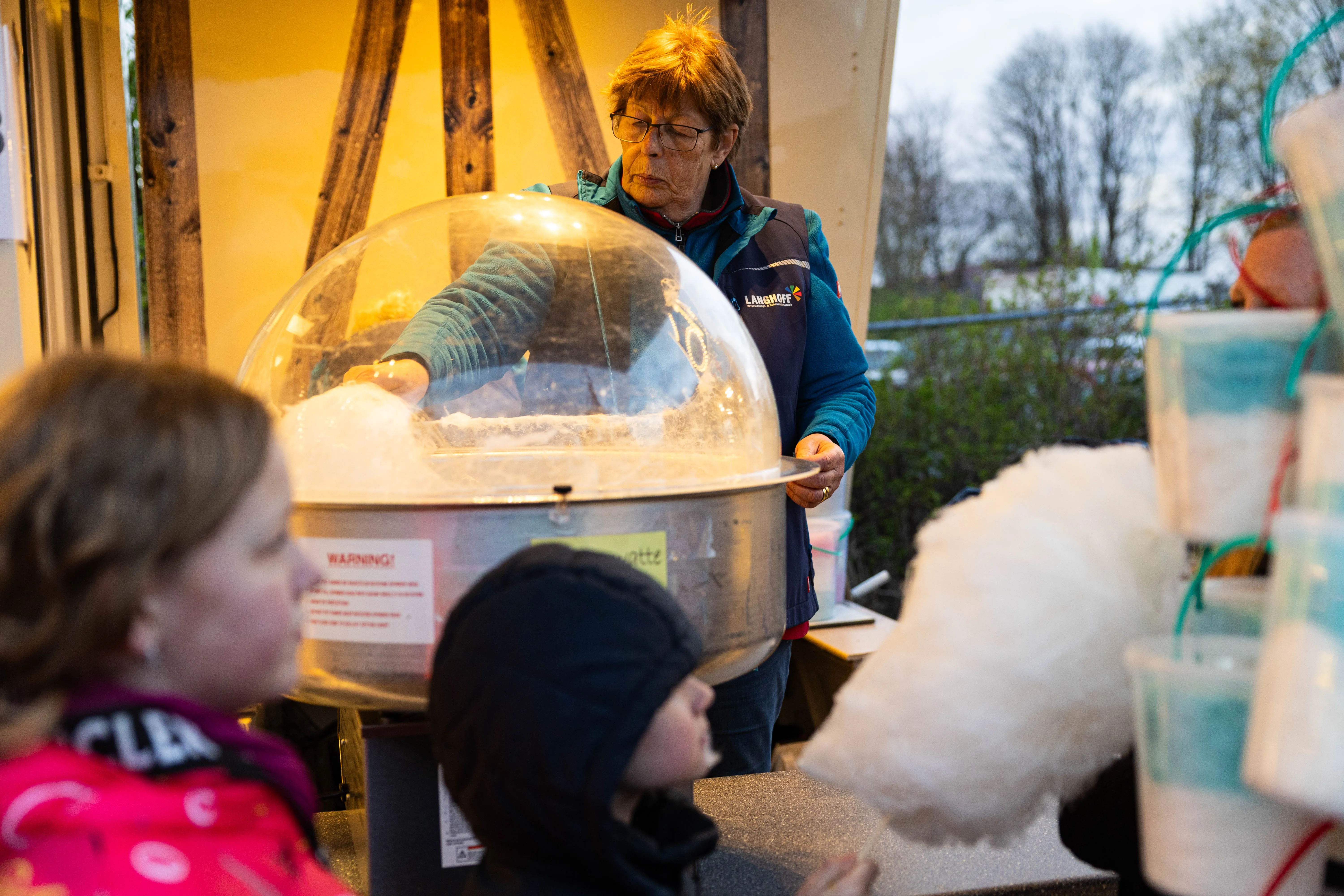 Eine Frau mit Brille und Weste steht an einer Zuckerwattemaschine, während Kinder vor ihr auf Zuckerwatte warten. Im Hintergrund ein Wohnwagen und Bäume.