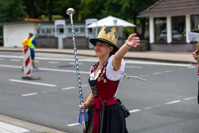 Eine Frau in traditioneller Tracht mit Krone und Hut hält einen verzierten Stab und hebt den Arm während einer Parade.
