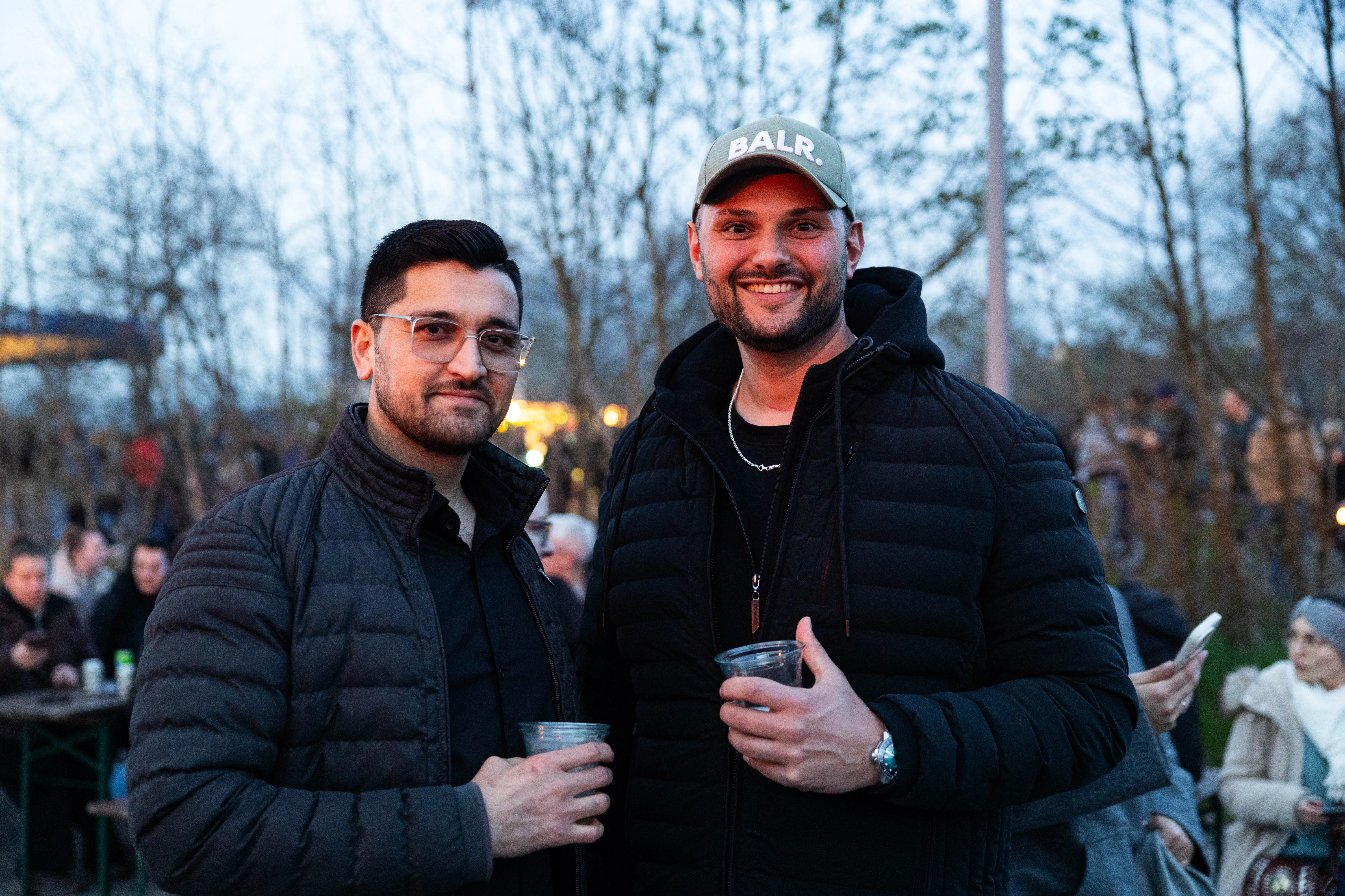 Zwei lächelnde Männer halten jeweils ein Glas in der Hand, im Hintergrund Menschen und Bäume bei einer Veranstaltung im Freien.