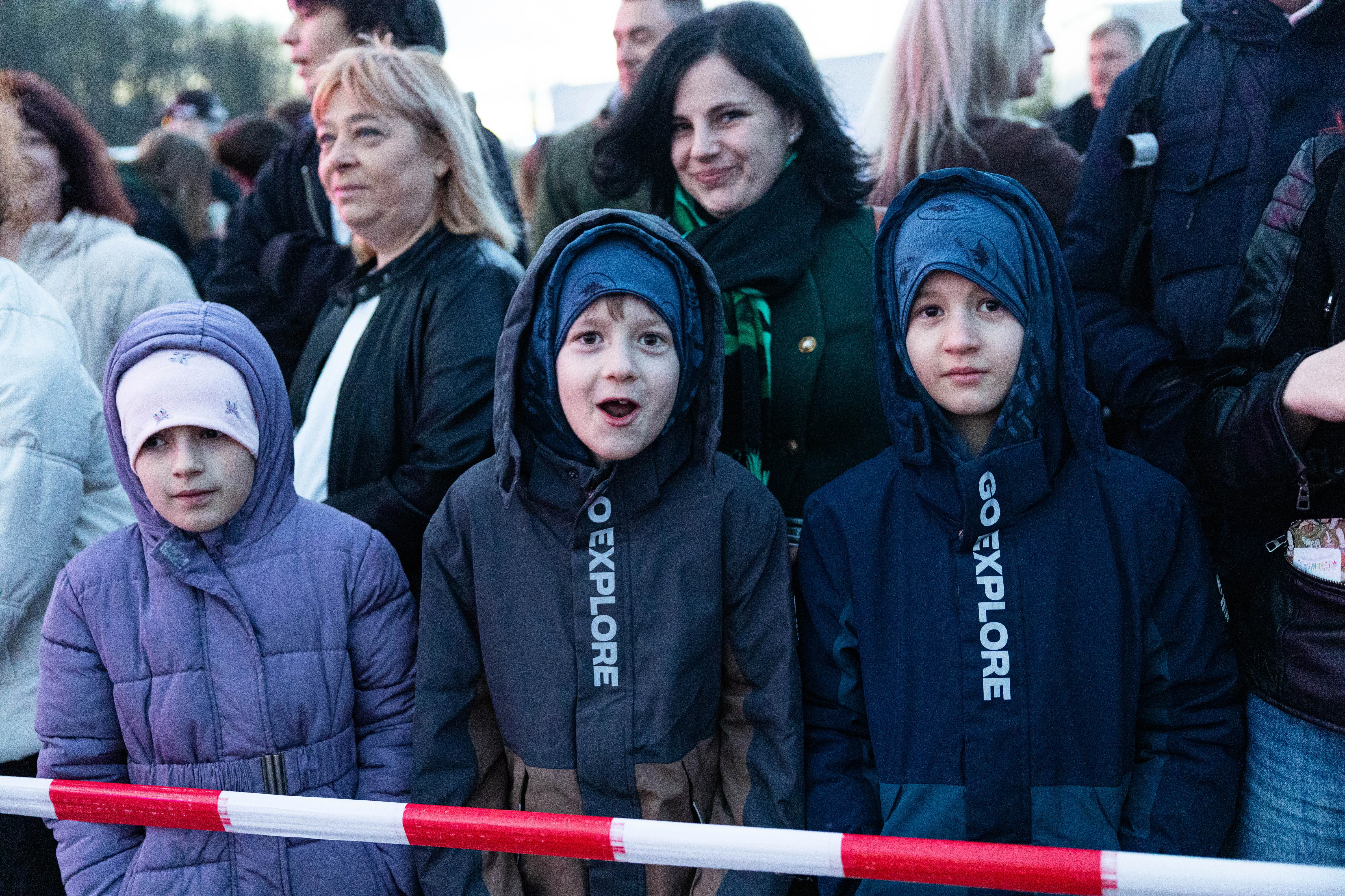 Drei Kinder mit Winterjacken und Kapuzen stehen vor einer Absperrung, im Hintergrund eine Menschenmenge. Die Jacken haben den Aufdruck „GO EXPLORE“.