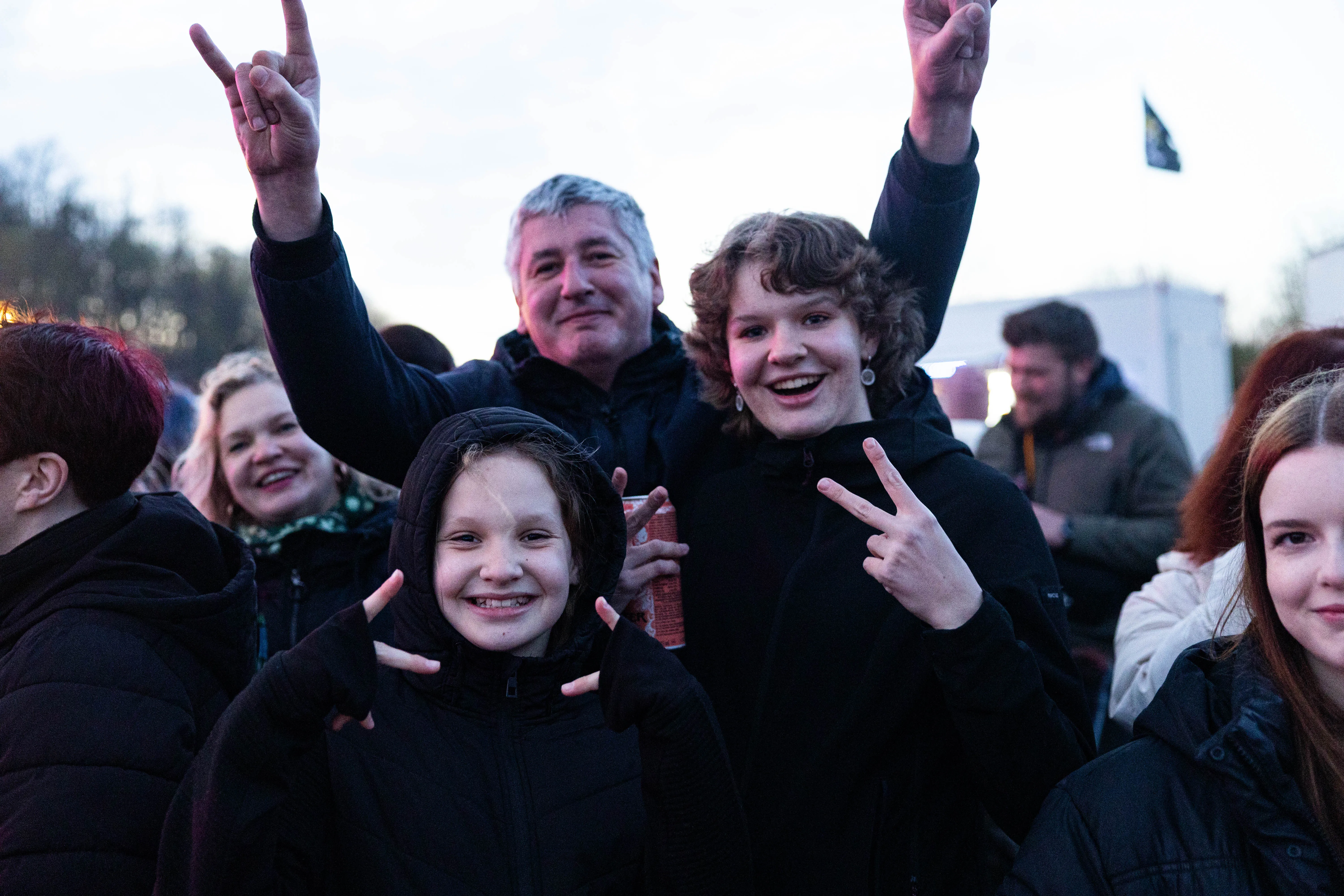 Mehrere Menschen lächeln in die Kamera und zeigen Handzeichen. Im Hintergrund sind weitere Zuschauer und ein Fahrzeug zu sehen.