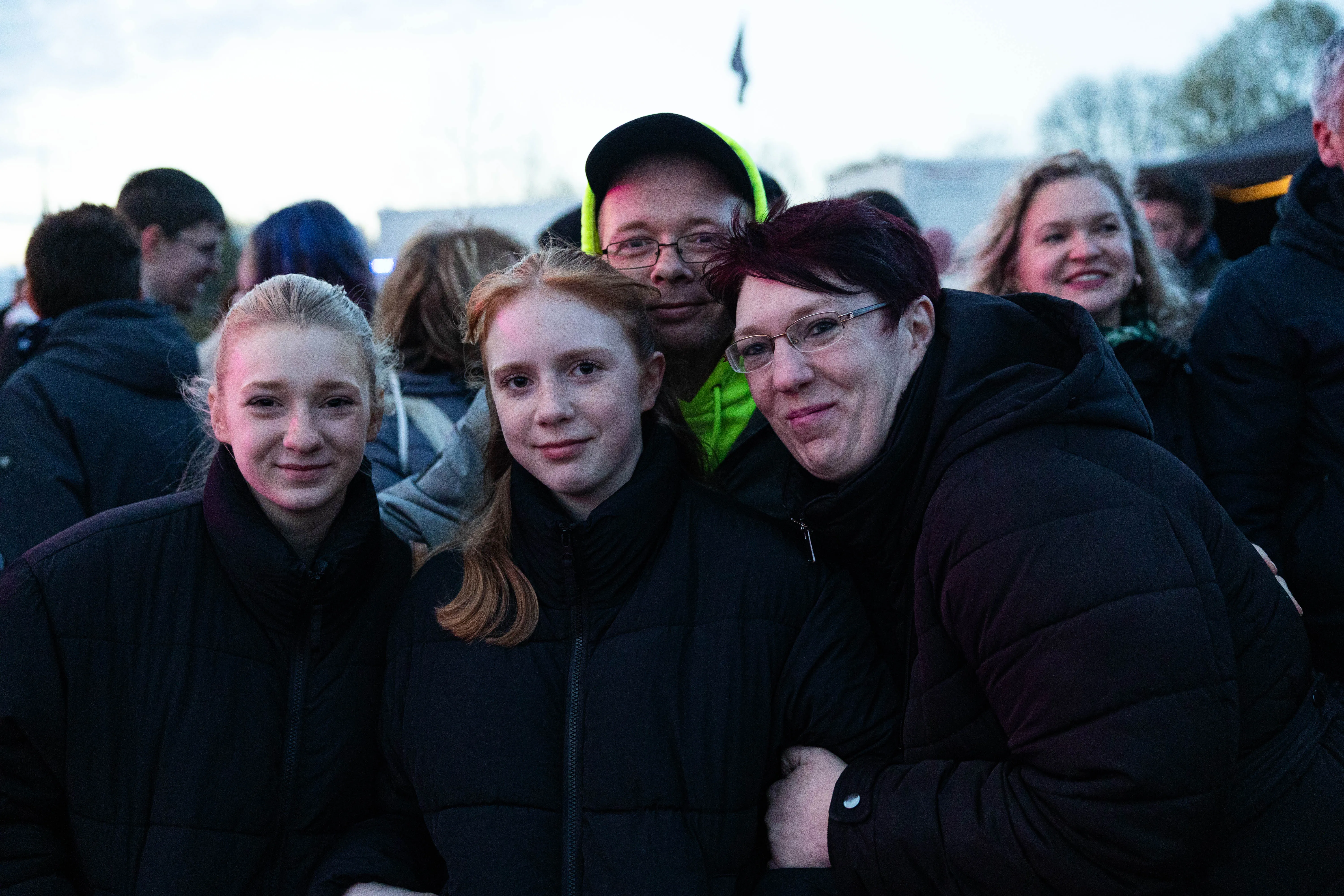 Drei Frauen und ein Mann lächeln in die Kamera, umgeben von einer Menschenmenge bei einer Veranstaltung im Freien.
