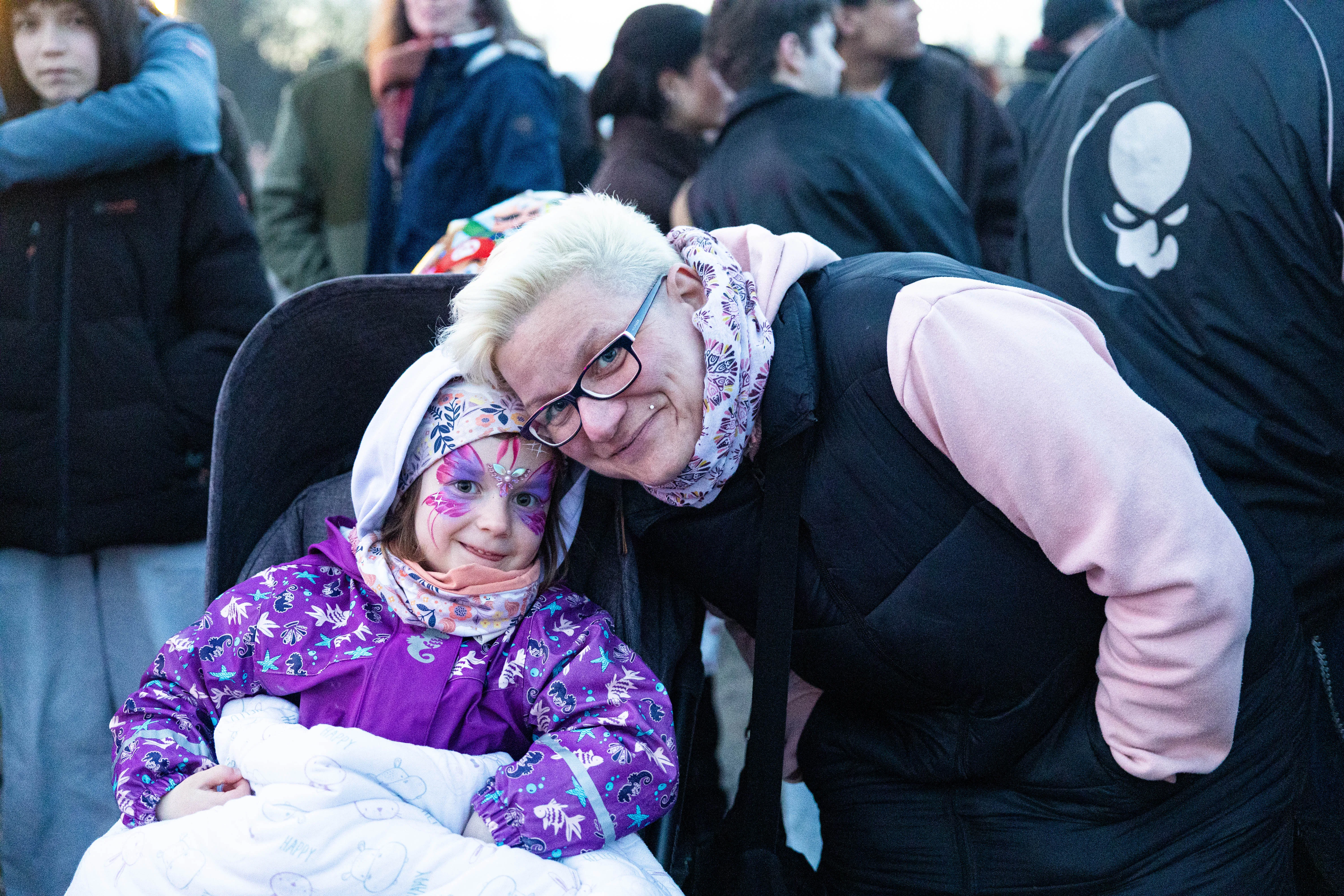 Eine Frau mit blonden Haaren umarmt ein kleines Mädchen im Rollstuhl. Im Hintergrund sind weitere Menschen zu sehen.