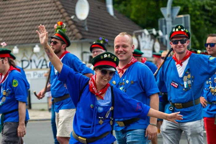 Eine lächelnde Frau in blauer Kleidung und grünem Hut winkt in die Kamera, umgeben von Mitgliedern der Meinerzhagener Schützengilde.