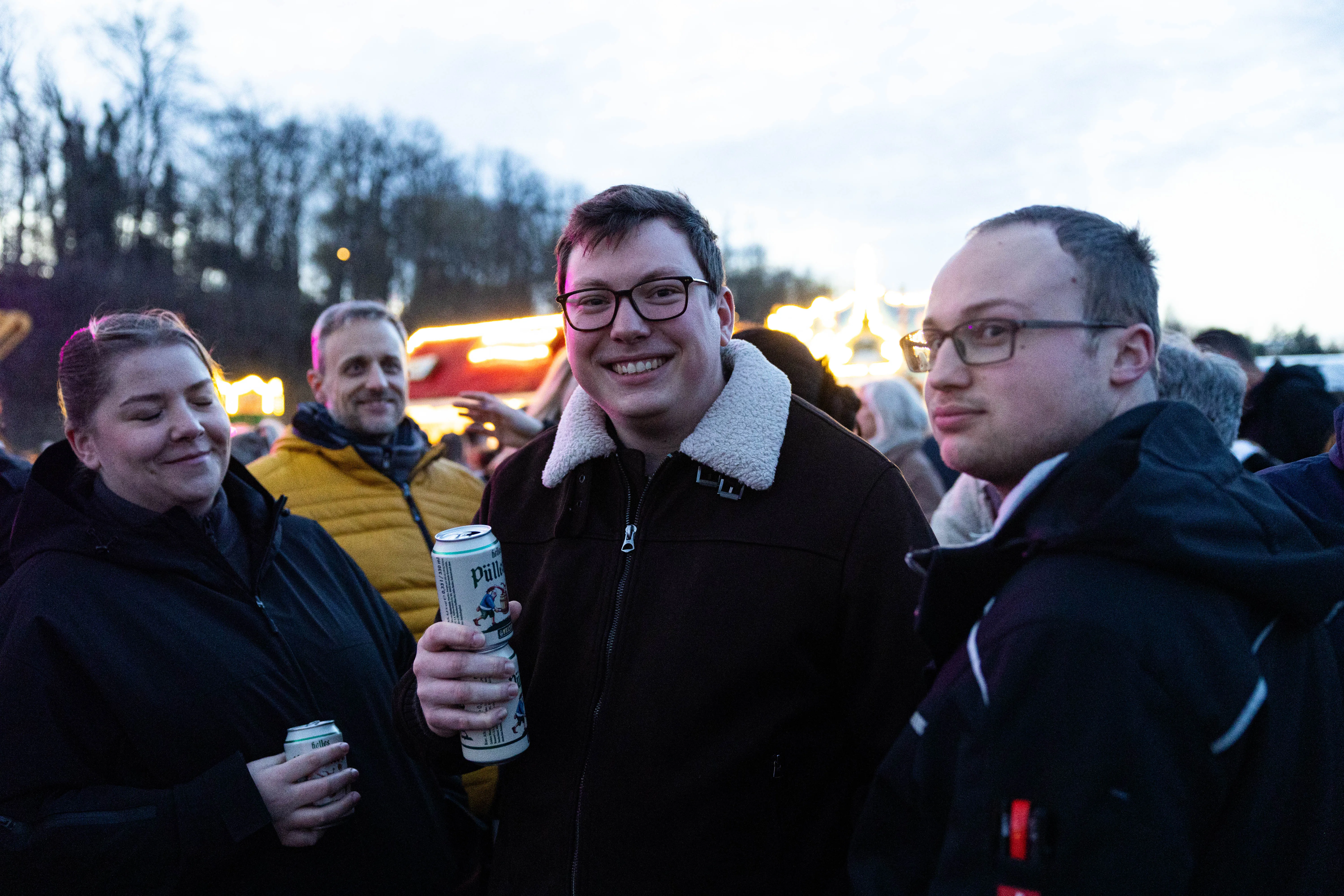 Drei junge Erwachsene lächeln in die Kamera, während sie auf einem Osterfeuer in Lüdenscheid stehen und Bierdosen halten. Im Hintergrund sind weitere Menschen und Feuerlichter zu sehen.