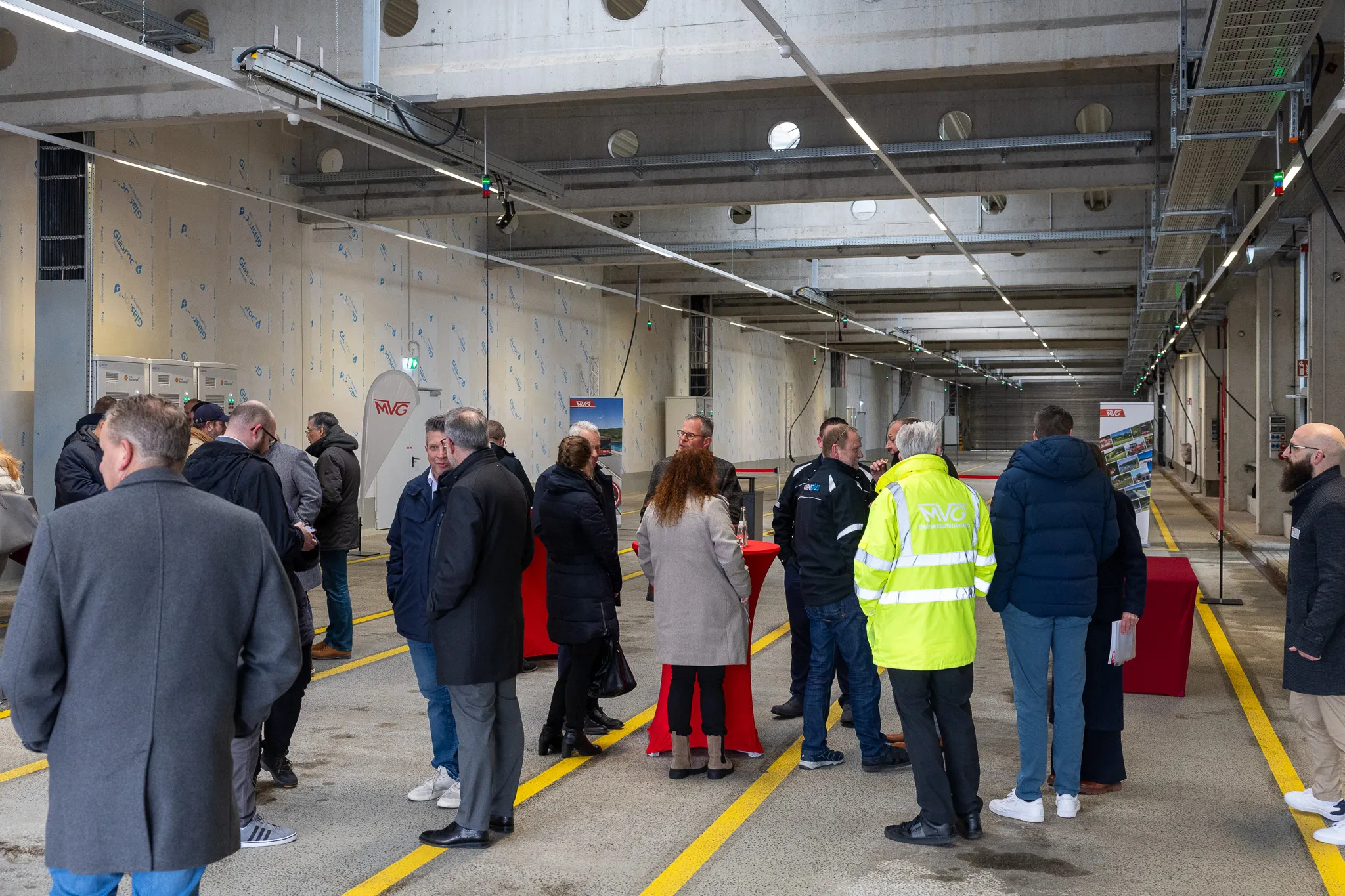 Gruppe von Menschen steht in einer modernen, hellen Halle mit Betonwänden und -decken. Im Hintergrund sind gelbe Markierungen auf dem Boden zu sehen und ein Tunnel führt in die Ferne. Mehrere Personen tragen Anzüge und Westen mit dem MVG-Logo.