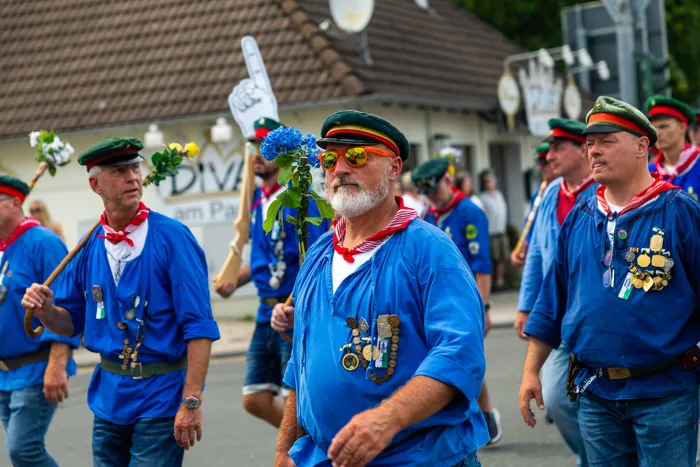 Stefan Schneider in blauem Hemd und grüner Kappe hält eine blaue Blume, während er mit Mitgliedern der Meinerzhagener Schützengilde durch die Straße geht.