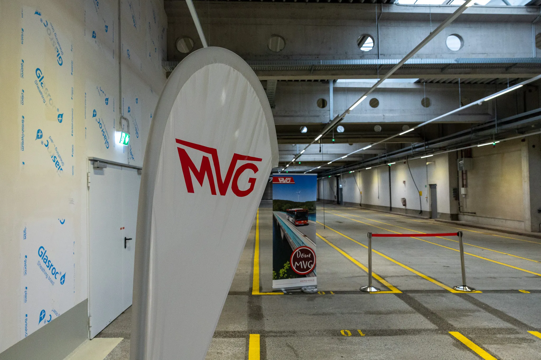 Großes weißes Banner mit rotem MVG-Logo steht vor einer Reihe von gelben Markierungen auf dem Boden einer modernen Bushalle.