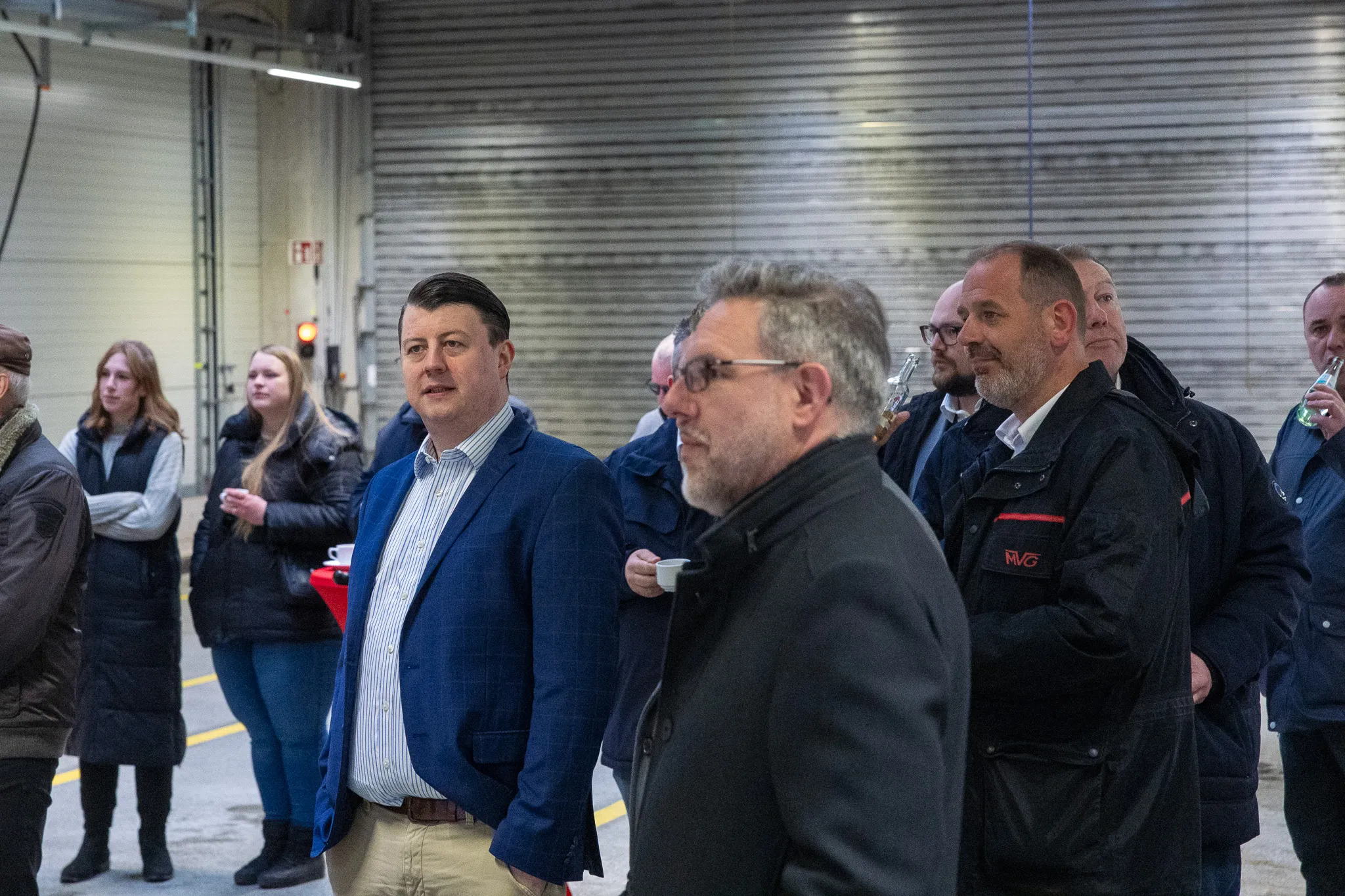 Mehrere Männer in Anzügen und Jacken stehen in einer großen Halle und blicken nach vorne, während im Hintergrund weitere Personen zu sehen sind. Im Vordergrund hält einer der Männer eine Flasche Wasser.
