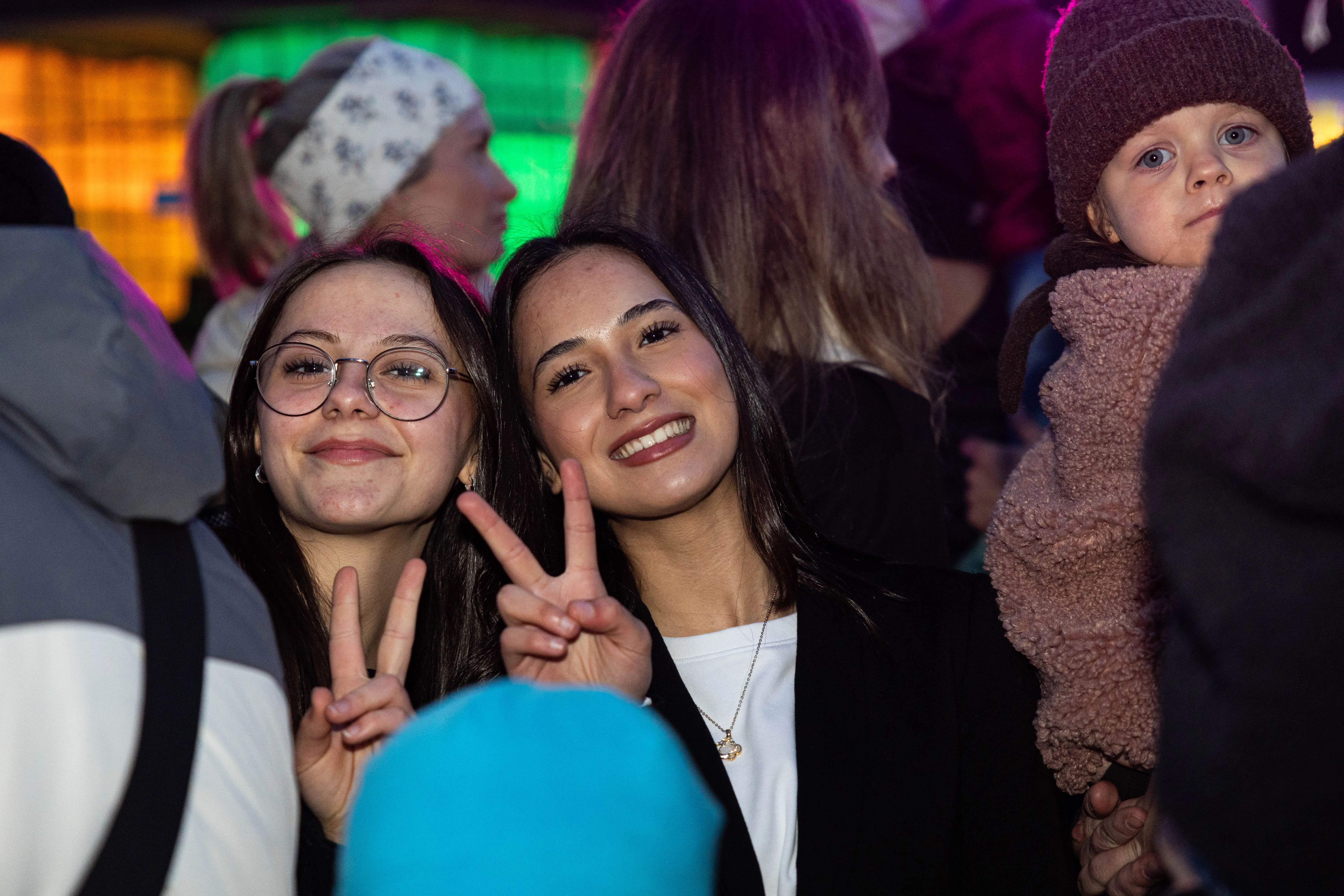 Zwei junge Frauen zeigen den Victory-Zeichen vor einer Menschenmenge bei einer Veranstaltung. Im Hintergrund ist ein Kind in einem braunen Wintermantel zu sehen.