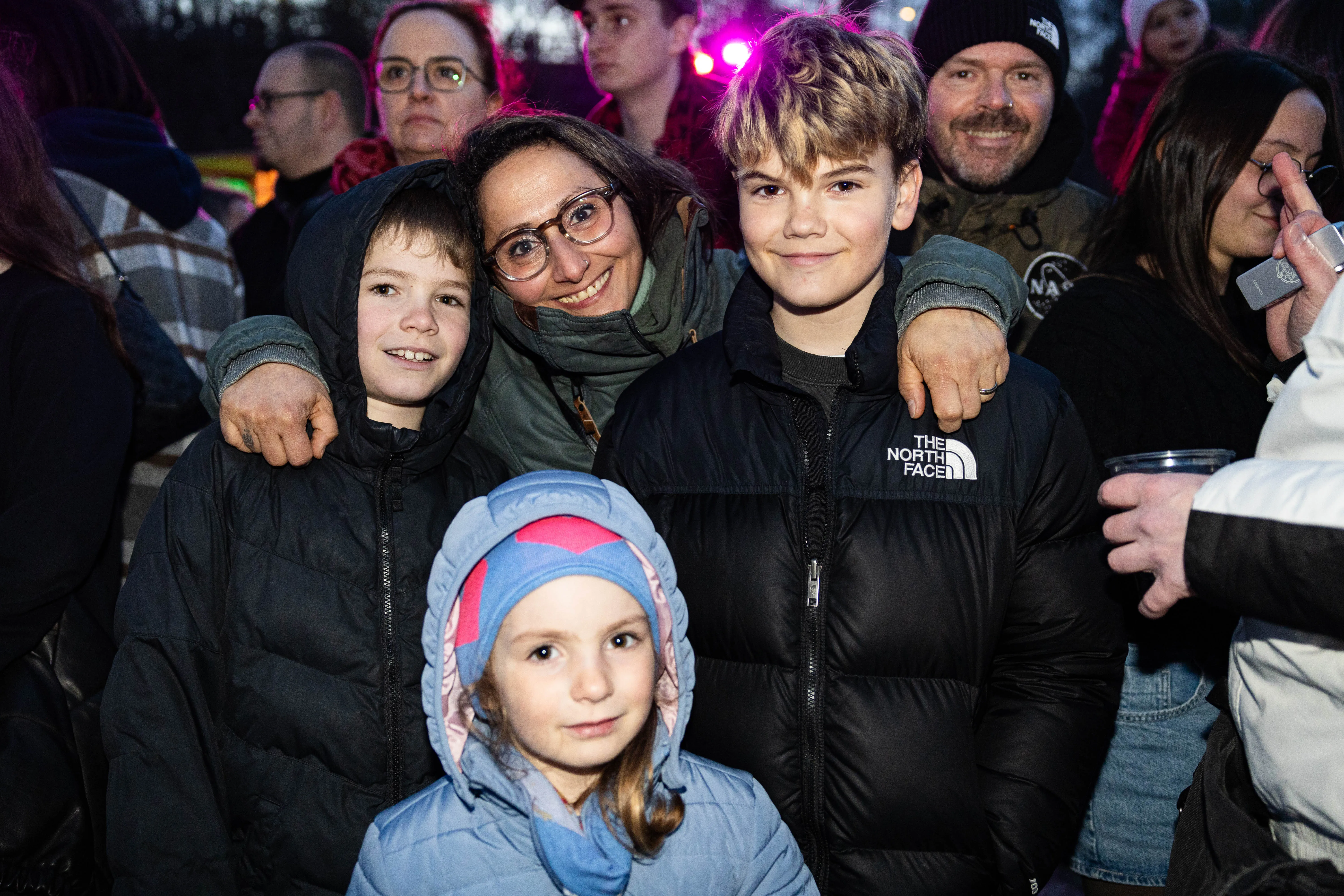 Eine Frau umarmt ihre beiden Kinder vor einer Menschenmenge bei einem Osterfeuer. Im Hintergrund sind weitere Menschen und Lichter zu sehen.