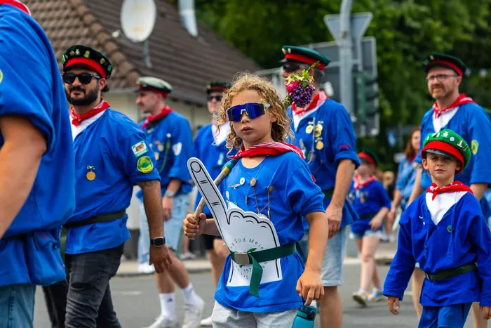 Ein Kind mit Sonnenbrille und einem großen Finger-Aufsteller läuft in einer Gruppe der Meinerzhagener Schützengilde in blauen Uniformen.