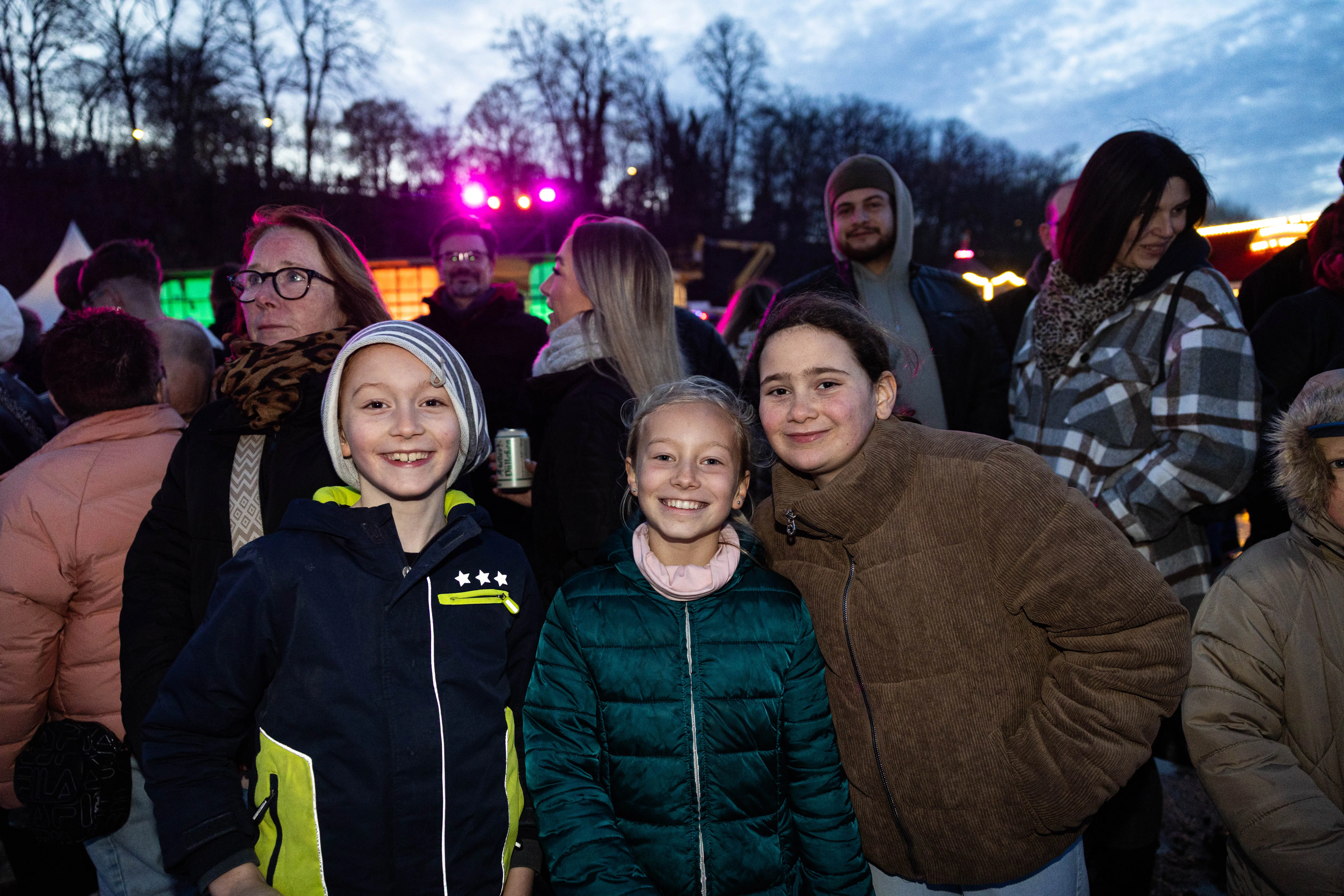 Drei lächelnde Kinder im Vordergrund, umgeben von einer Menschenmenge bei einer Abendveranstaltung mit Lichtern und Bäumen im Hintergrund.