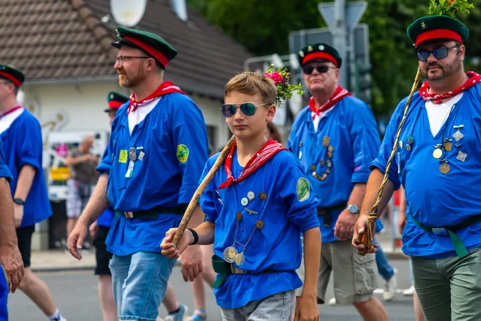 Mitglieder der Meinerzhagener Schützengilde in blauen Hemden und roten Halstüchern ziehen in einer Prozession. Ein Junge mit Sonnenbrille geht voran.