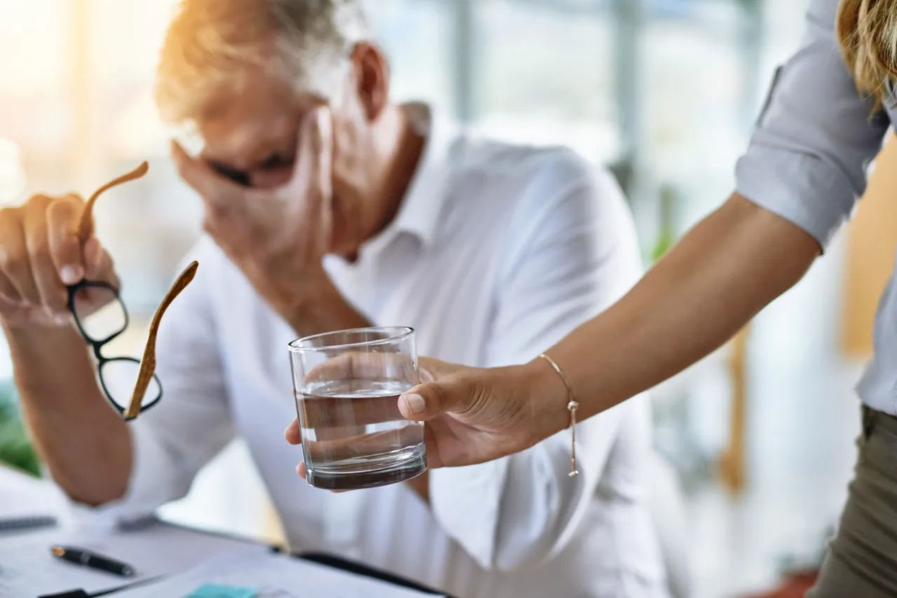 Ein Mann hält sich mit der Hand die Stirn, während ihm eine andere Person ein Glas Wasser reicht. Im Hintergrund liegen Dokumente auf einem Schreibtisch.