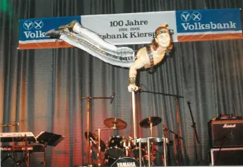 Künstler in weißer Kleidung hält sich an einer Stange über einer Schlagzeuganlage, im Hintergrund ein Banner mit dem Schriftzug „100 Jahre Volksbank Kierspe 1901-2001“.