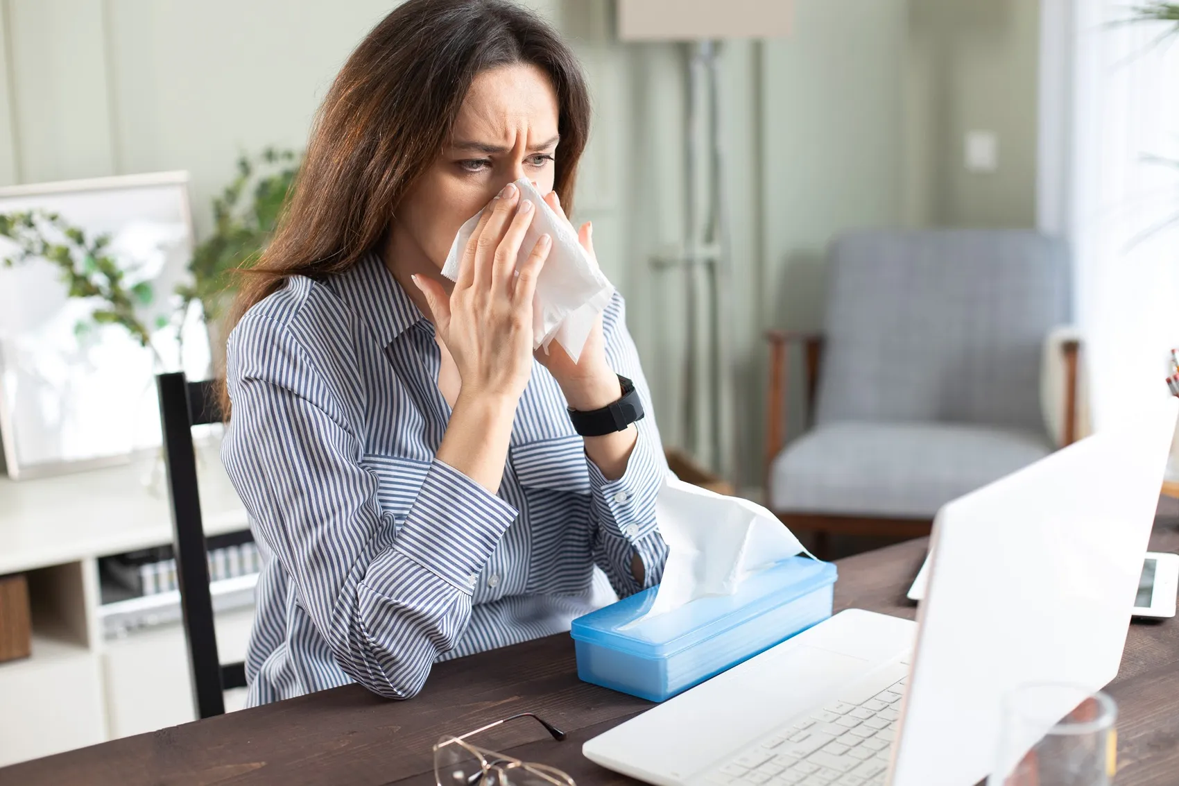 Eine Frau hält sich ein Taschentuch vor die Nase und schaut angespannt auf einen geöffneten Laptop. Vor ihr steht eine Packung Taschentücher.
