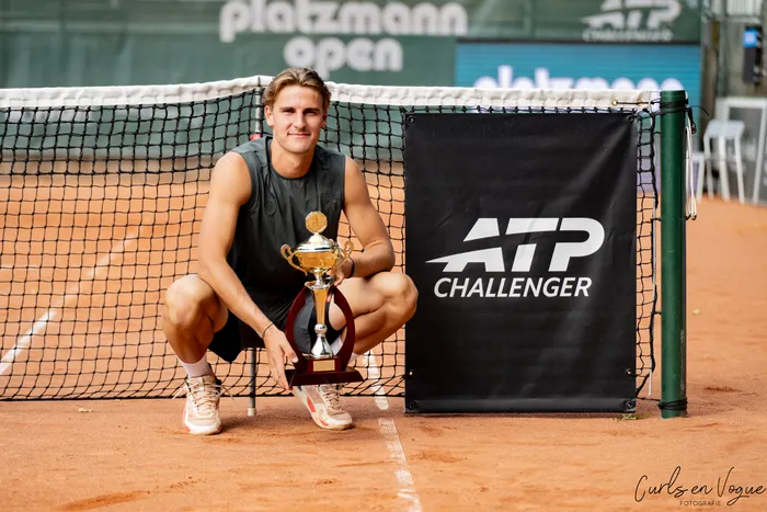 Tom Gentzsch hockt auf einem Sandplatz hinter einem Tennisnetz und hält eine goldene Trophäe neben einem Schild mit der Aufschrift „ATP Challenger“.