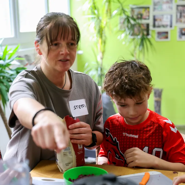 Eine Frau mit kurzen, dunklen Haaren zeigt einem Jungen in rotem Trikot eine Packung Lebensmittel. Sie stehen an einem Tisch mit Zutaten und Schüsseln, im Hintergrund sind Pflanzen und Bilder an der Wand zu sehen.