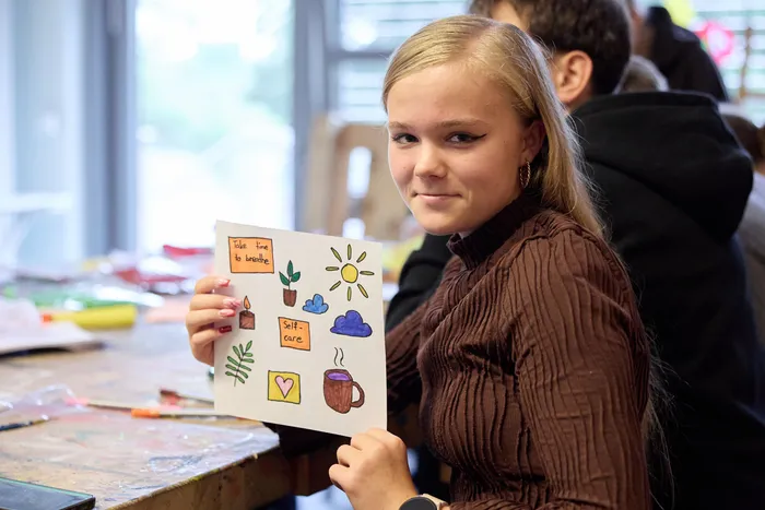 Eine junge Frau hält ein farbiges, handgezeichnetes Plakat mit Motiven zu Selbstfürsorge und Wohlbefinden hoch. Darauf sind Pflanzen, eine Sonne, eine Tasse und der Schriftzug „Self-care“ zu sehen.