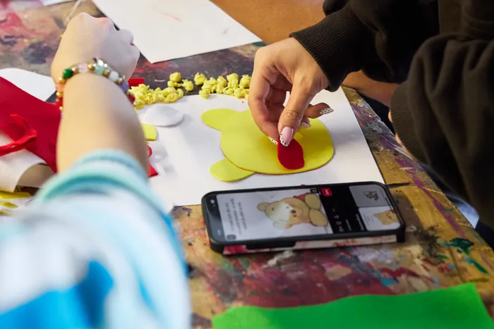 Hände formen aus gelbem und rotem Fondant einen Bären auf einem Tisch, während ein Smartphone mit einem Bild des Bären darauf im Vordergrund liegt. Das Bild zeigt die kreative Arbeit von Jugendlichen im Rahmen eines deutsch-polnischen Jugendprojekts zum Thema Wohlbefinden.