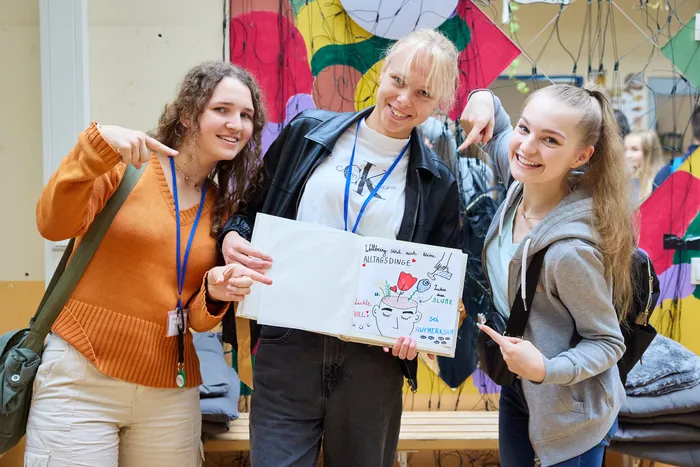 Vier junge Menschen, drei Frauen und ein Mann, halten ein Plakat mit Zeichnungen und Texten zum Thema Wohlbefinden. Im Hintergrund eine bunte Wand und Kleidung.
