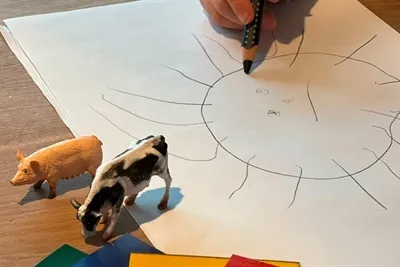 Eine Hand zeichnet eine Sonne auf ein weißes Blatt Papier, neben dem eine Spielzeugsau und eine Kuh stehen.