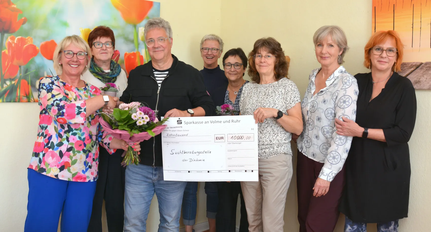 Acht Personen posieren mit einem Blumenstrauß und einem symbolischen Scheck über 10.000 Euro für die Suchtberatungsstelle der Diakonie.