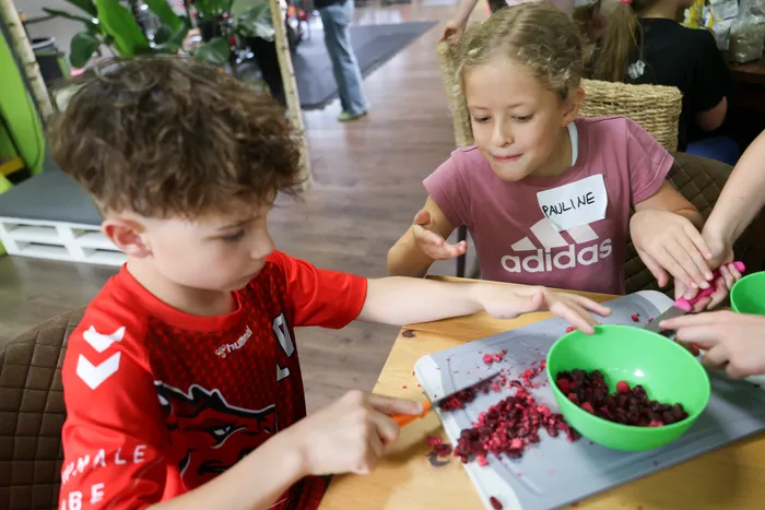 Zwei Kinder bereiten gemeinsam Beeren auf einem Schneidebrett zu. Der Junge trägt ein rotes Trikot, das Mädchen ein rosafarbenes T-Shirt mit dem Namensschild 