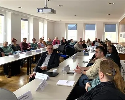 Verschiedene Personen sitzen an Tischen in einem hellen Konferenzraum zusammen und diskutieren. Ein Mann im Anzug blickt auf sein Smartphone.
