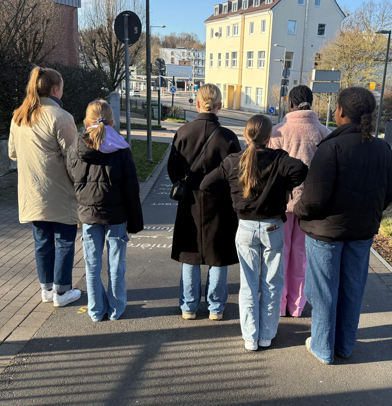 Eine Gruppe junger Frauen steht mit dem Rücken zum Betrachter auf einem Gehweg und blickt in eine Richtung. Im Hintergrund sind Gebäude und Bäume zu sehen.