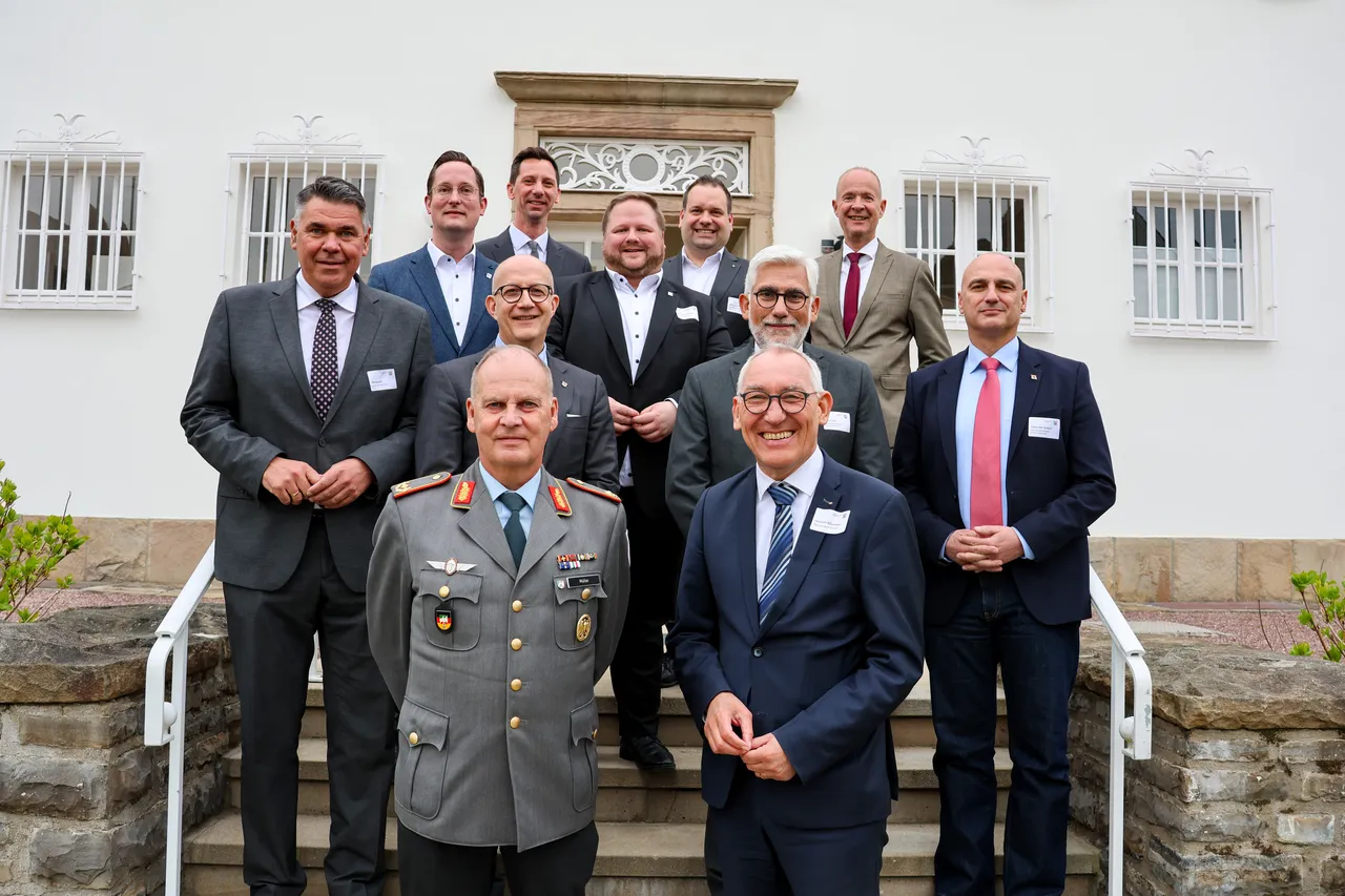 Gruppe von Männern in Anzügen posiert auf einer Treppe vor einem hellen Gebäude. Im Vordergrund stehen zwei Männer in Uniformen, während die anderen in Businesskleidung sind.