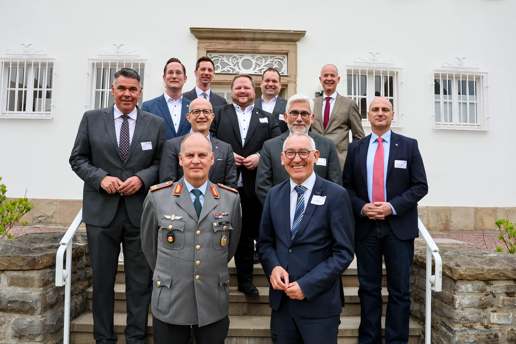 Gruppe von Männern in Anzügen posiert auf einer Treppe vor einem hellen Gebäude. Im Vordergrund stehen zwei Männer in Uniformen, während die anderen in Businesskleidung sind.