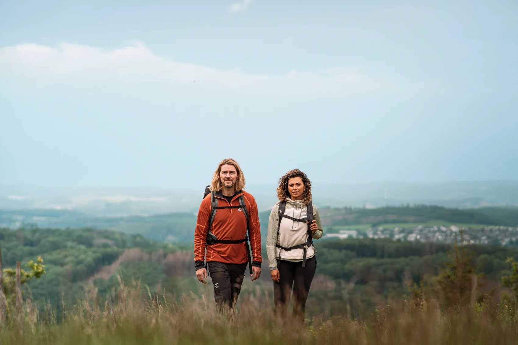 Zwei Wanderer gehen auf einem grasbewachsenen Hügelrücken, im Hintergrund eine hügelige Landschaft mit Wald und Ortschaft.