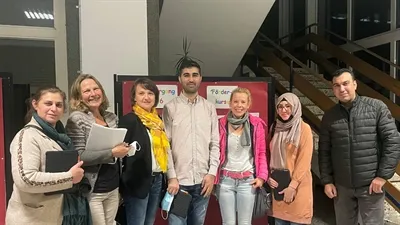 Gruppe von sieben Personen steht vor einer Treppe und einem roten Plakat mit Aufschriften in verschiedenen Sprachen. Sie halten Unterlagen und Taschen bei sich.