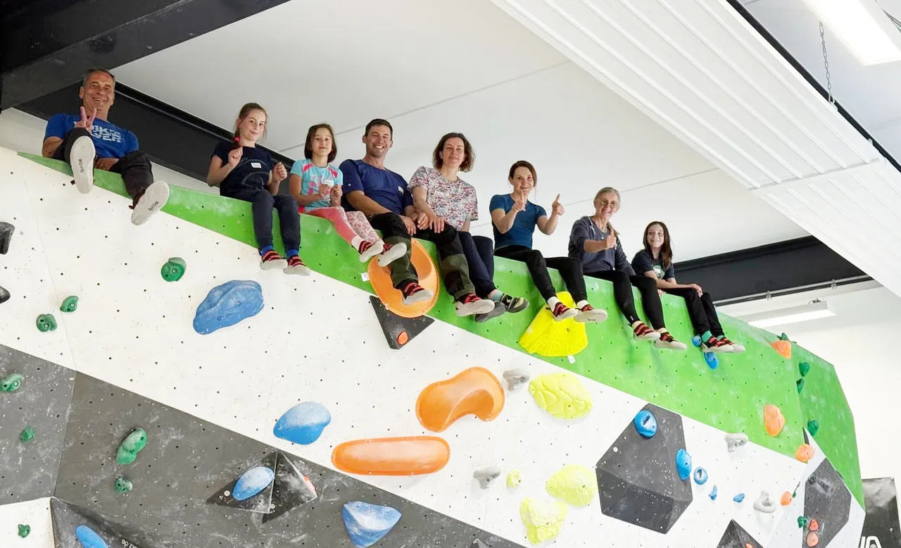 Eine Gruppe von sieben Personen sitzt auf bunten Griffen einer Kletterwand. Sie lächeln in die Kamera und wirken motiviert.