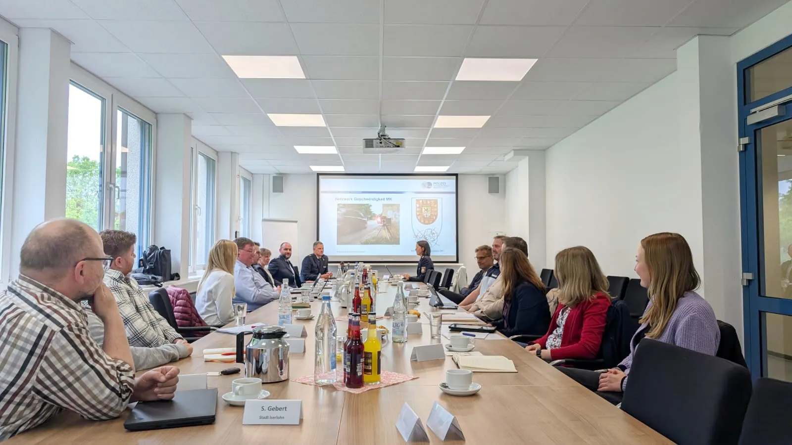 Verschiedene Personen sitzen an einem Konferenztisch und blicken auf eine Präsentation über Verkehrssicherheit, die eine Straße und ein Wappen zeigt. Im Vordergrund steht ein Namensschild mit der Aufschrift 'S. Gebert'.