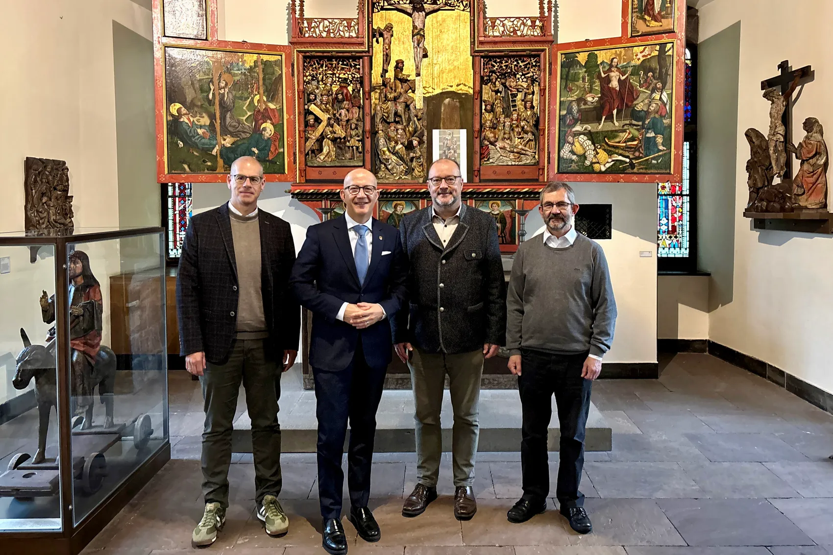 Vier Männer stehen in einem Raum mit mittelalterlichen Kunstwerken vor einem großen Altar. Von links nach rechts sind es Oliver Günther, Landrat Ralf Schwarzkopf, Dr. Christof Grote und Claus Optenhöfel.