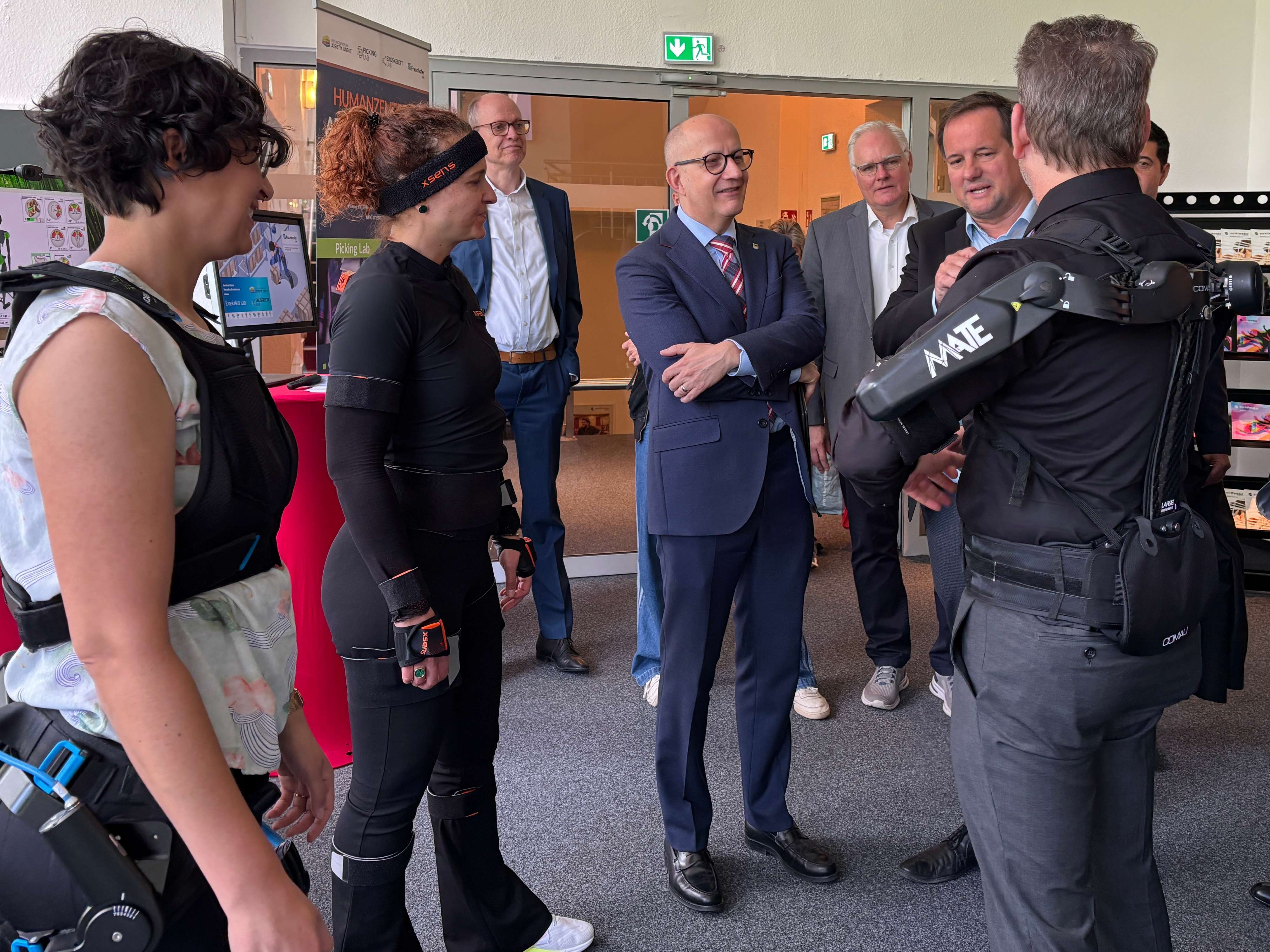 Zwei Männer in Anzügen und zwei Frauen, eine mit einer Exoskelett-artigen Vorrichtung, stehen in einer Messehalle und unterhalten sich. Im Hintergrund sind weitere Stände und Besucher zu sehen.