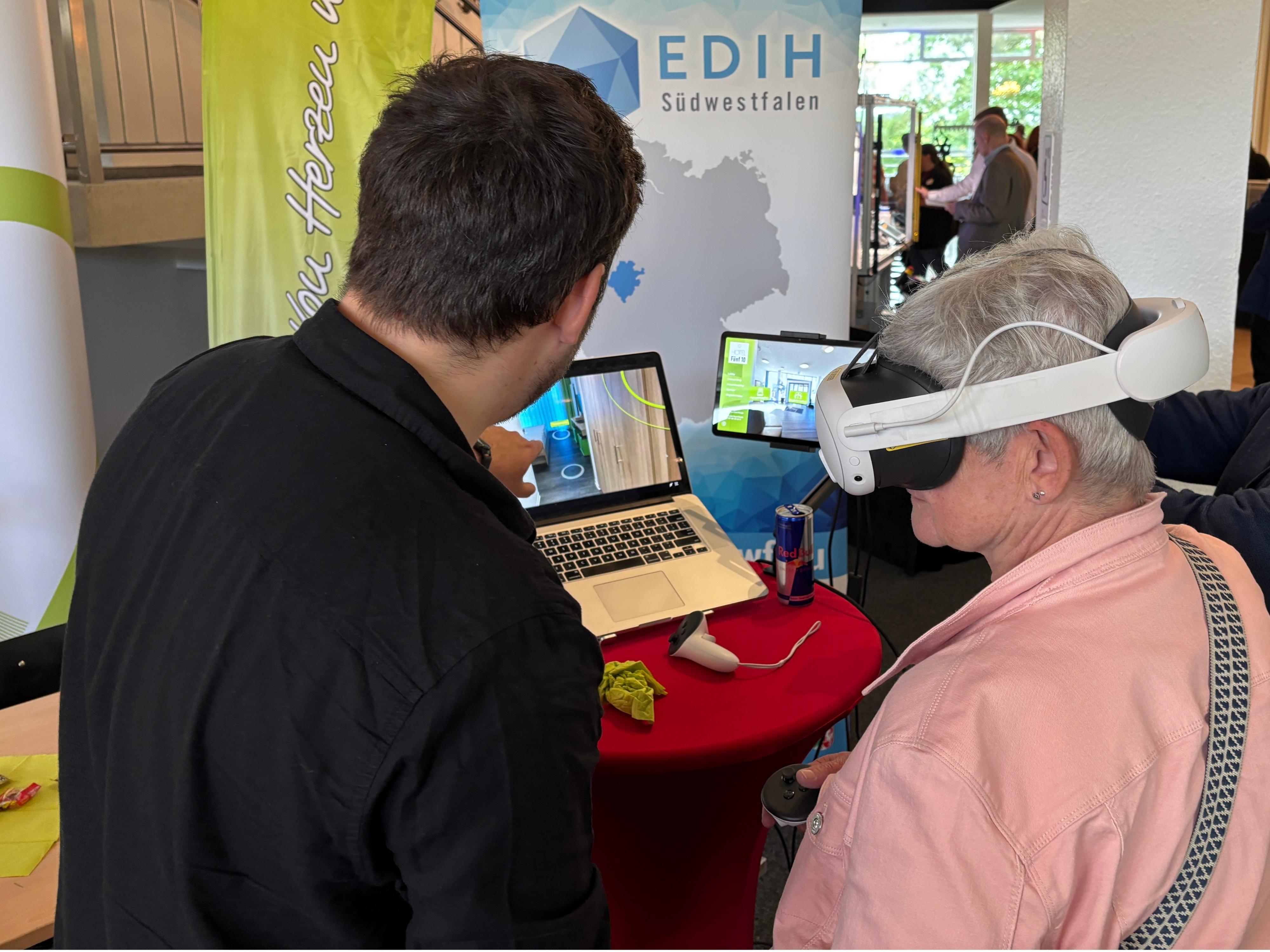 Ein Mann erklärt einer älteren Frau mit VR-Brille die Bedienung einer virtuellen Hotelumgebung auf einem Laptop.