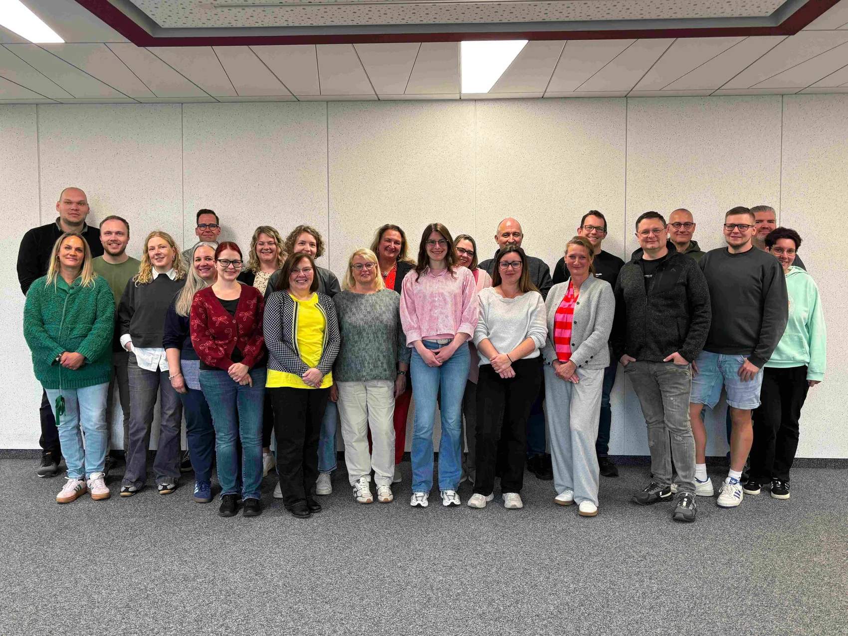 Gruppe von 26 Fachkräften aus dem Gesundheitswesen steht für ein gemeinsames Foto in einem hellen Raum zusammen.