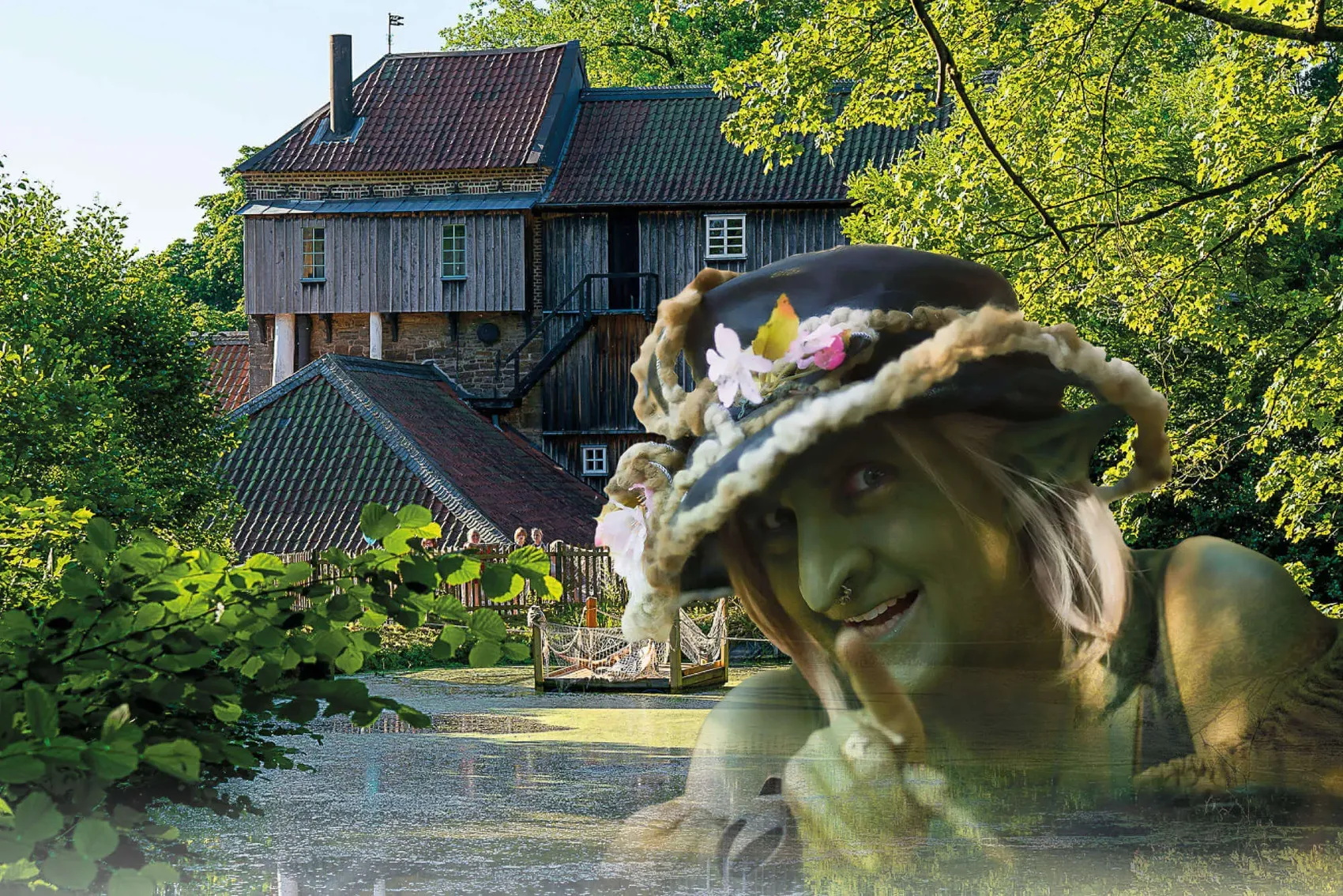 Eine Frau mit Blumen geschmücktem Hut lächelt aus dem Wasser, im Hintergrund eine historische Mühle und Besucher auf einer Brücke.