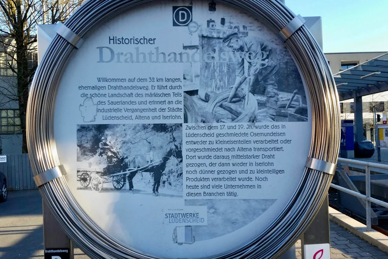 Historisches Schild des Drahthandelswegs zeigt eine Schwarz-Weiß-Fotografie von Arbeitern mit Werkzeugen vor einer Schmiede. Der Text beschreibt die industrielle Vergangenheit der Städte Lüdenscheid, Altena und Iserlohn und die Verarbeitung von Draht zwischen dem 17. und 19. Jahrhundert.