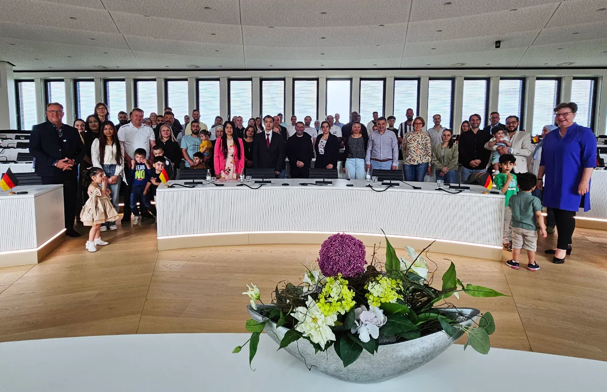 Eine Gruppe von 62 Menschen steht im Kreishaus Lüdenscheid zusammen, nachdem sie die deutsche Staatsbürgerschaft erhalten haben. Im Vordergrund steht ein Blumenarrangement.