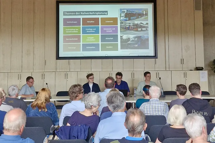 Eine Gruppe von Menschen sitzt um einen Konferenztisch, während ein Mann an der Stirnseite spricht. Im Hintergrund ist eine Präsentation über Nahverkehrsplanung mit Bildern von Bussen und Bahnen zu sehen.
