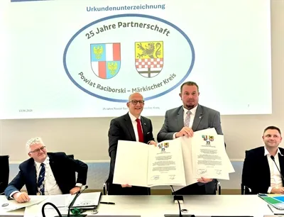 Zwei Männer in Anzügen halten ein geöffnetes Dokument mit Unterschriften vor sich. Im Hintergrund ein Banner mit den Wappen von Powiat Raciborski und Märkischer Kreis sowie der Aufschrift '25 Jahre Partnerschaft'.