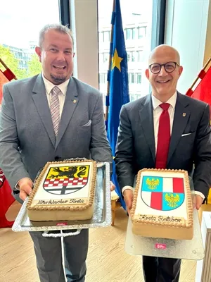 Zwei Männer in Anzügen halten jeweils eine rechteckige Torte, die mit einem Wappen verziert ist. Die Torte links zeigt einen schwarzen Adler auf rot-weißem Schachbrettmuster, die rechte einen roten Adler auf weißem Grund.