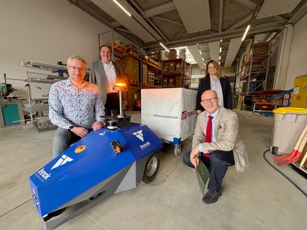 Four people stand in a warehouse with a blue robot in the foreground. From left to right are Nico Hackmann, Jochen Schröder, Sonja Pfaff, and Ralf Schwarzkopf. The robot has 