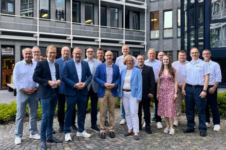 Eine Gruppe von etwa 15 Personen, überwiegend Männer in Anzügen, steht lächelnd vor einem modernen Gebäude mit grauer Fassade. Sie nehmen an einer Informationsveranstaltung des Märkischen Kreises für Hauptverwaltungsbeamte teil.